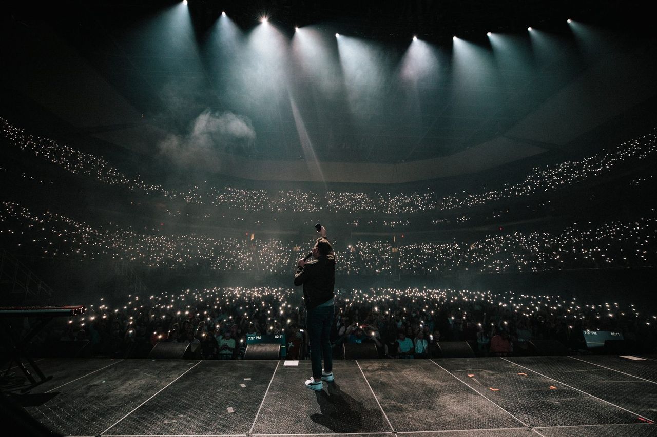 Wide shot from behind the stage of a full arena lit by phone lights at Elevation Nights, Austin