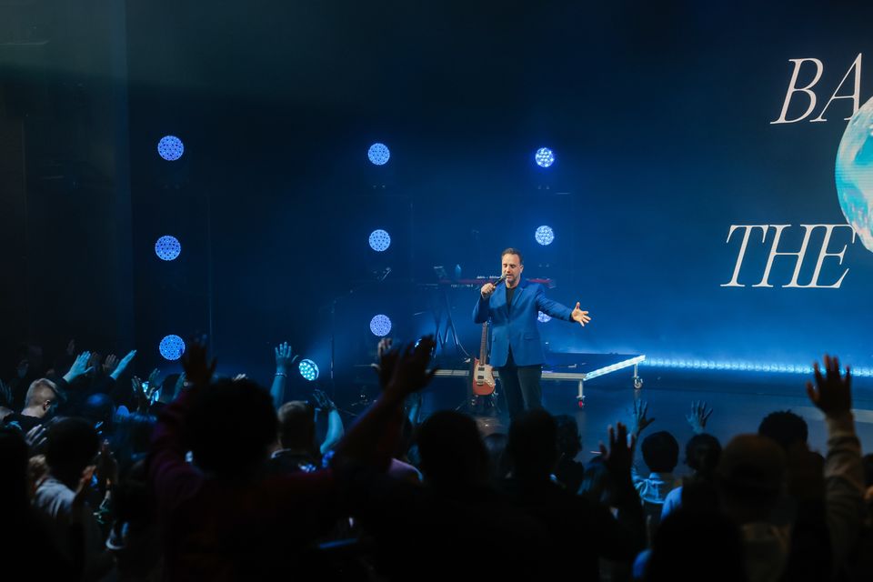 Mark Francey on stage under the blue Baptize the World globe, hands raised across the theater
