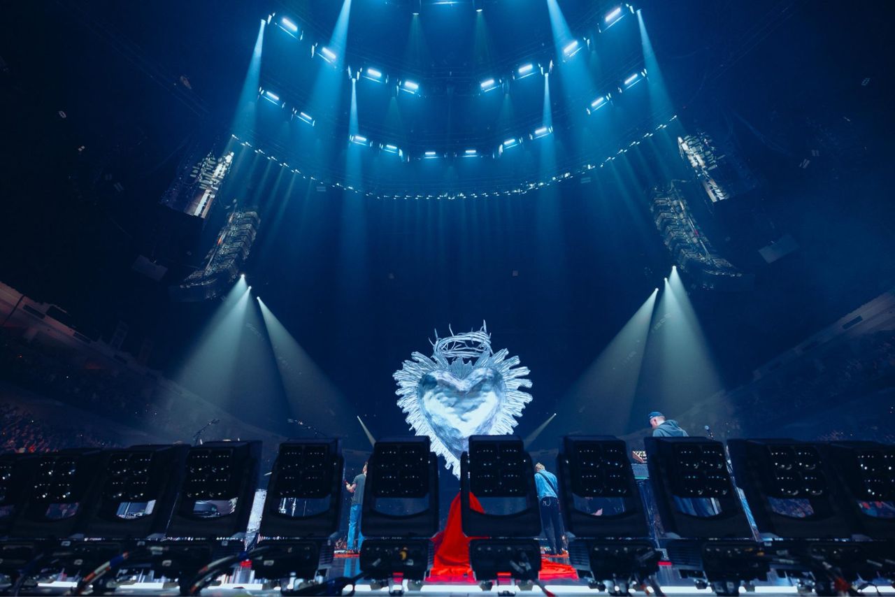 Wide from behind the stage across a full arena lit by phone lights