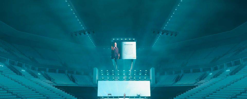 Design render of the preaching look — the cross-shaped stage and screens in teal light from high in the bowl